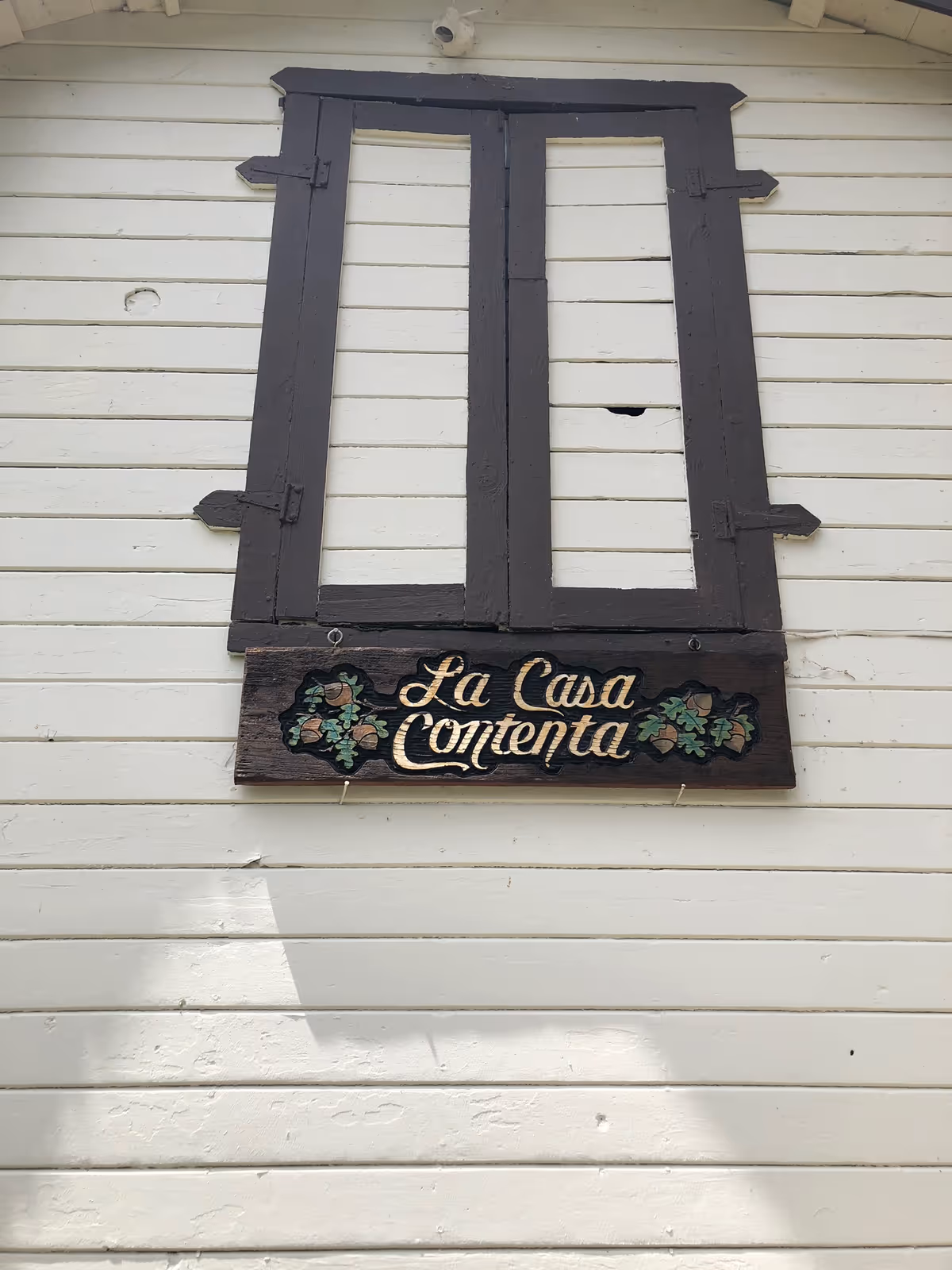 A white wooden exterior wall with a closed dark brown wooden window shutter. Below the window is a wooden sign with decorative leaves on each side and the text 'La Casa Contenta' painted in gold.