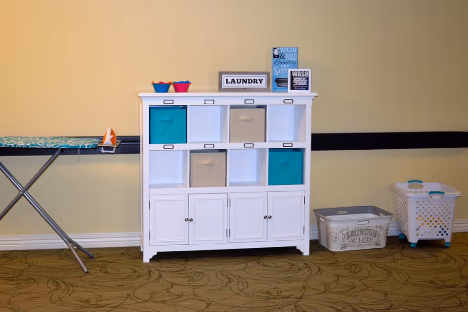 A laundry area with a white storage cabinet containing colorful bins, a small ironing board with an iron on the left, and two laundry baskets on the floor to the right. The wall behind is painted yellow with a black horizontal stripe.
