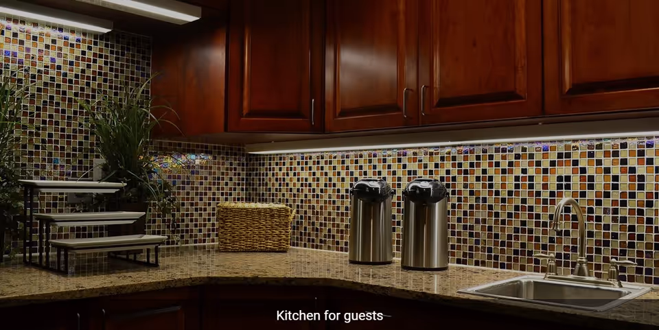 A guest kitchen area with dark wooden cabinets, a granite countertop, a stainless steel sink with a faucet, two stainless steel airpots, a woven basket, a tiered display stand, and a colorful mosaic tile backsplash.