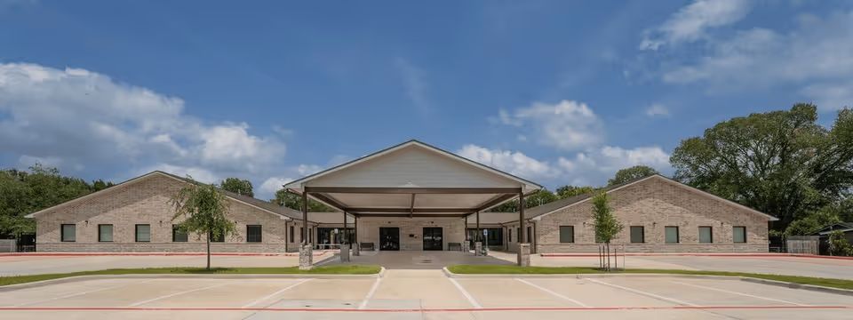 Front exterior view of Precisive Care I and II facility showing a single-story building with a covered entrance, surrounded by a parking lot and some small trees under a partly cloudy sky.