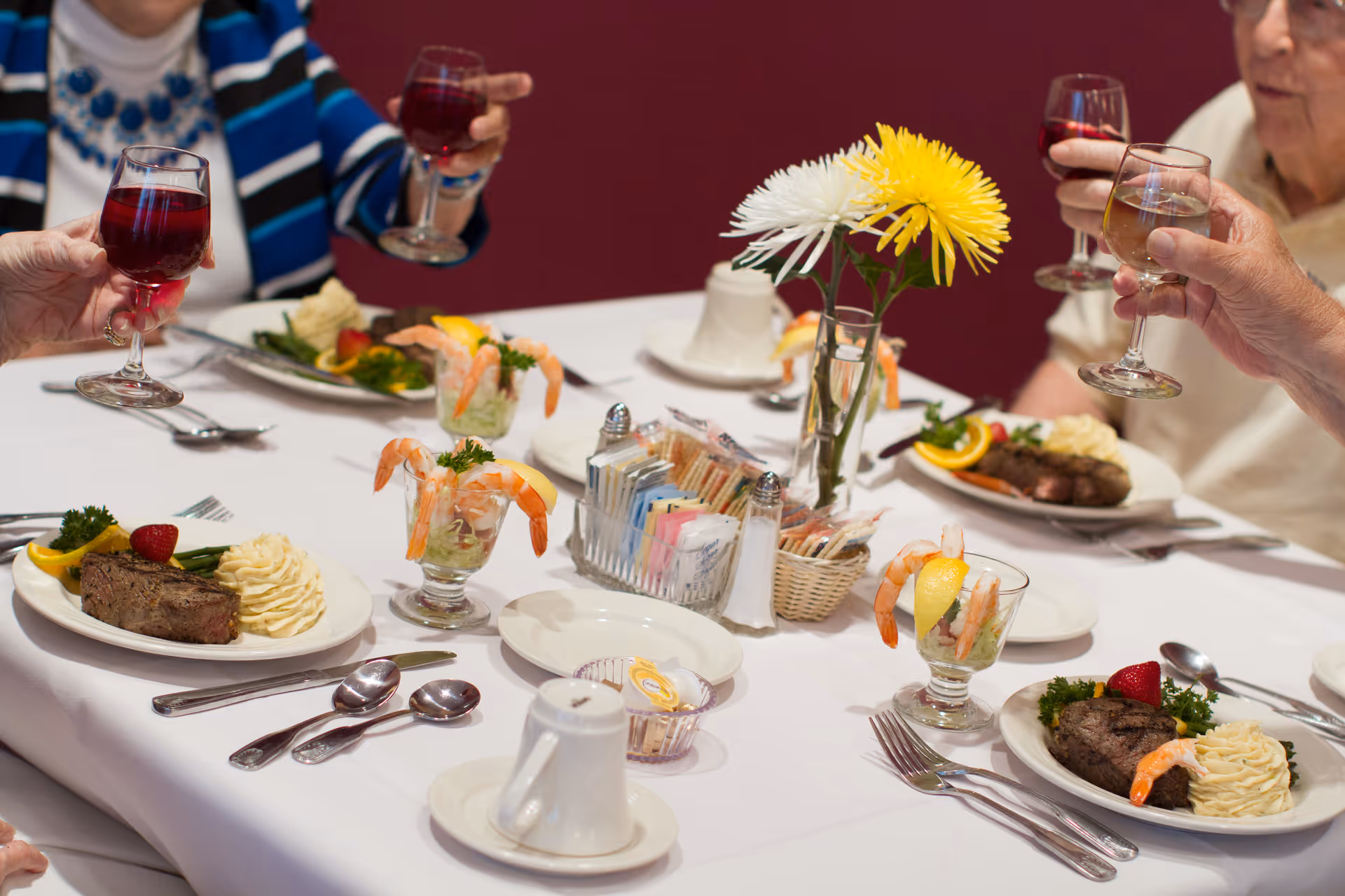 A group of elderly people sitting around a dining table raising glasses of red and white wine in a toast. The table is set with plates of steak, mashed potatoes, green beans, shrimp cocktail, and garnished with lemon and parsley. A vase with yellow and white flowers and a condiment holder with salt, pepper, and sugar packets are in the center of the table.