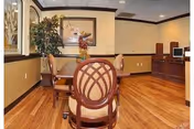 A small seating area with a round wooden table and four upholstered chairs on a polished wooden floor. The walls are decorated with framed artwork and a large potted plant is placed in the corner. There is a wooden cabinet with a computer monitor on the right side of the room.