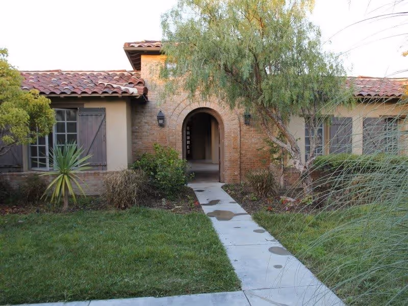 Exterior view of a single-story building with a tiled roof and an arched entrance. There is a concrete pathway leading to the entrance, surrounded by green grass, bushes, and trees.