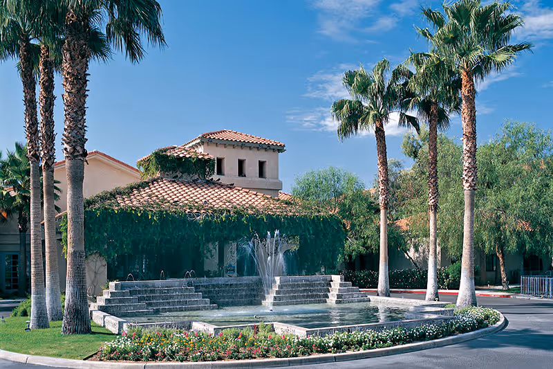 Exterior view of a building with a red-tiled roof partially covered in green vines, surrounded by tall palm trees and other greenery. In front of the building is a tiered stone fountain with water jets, bordered by a circular flower bed with colorful flowers under a clear blue sky.