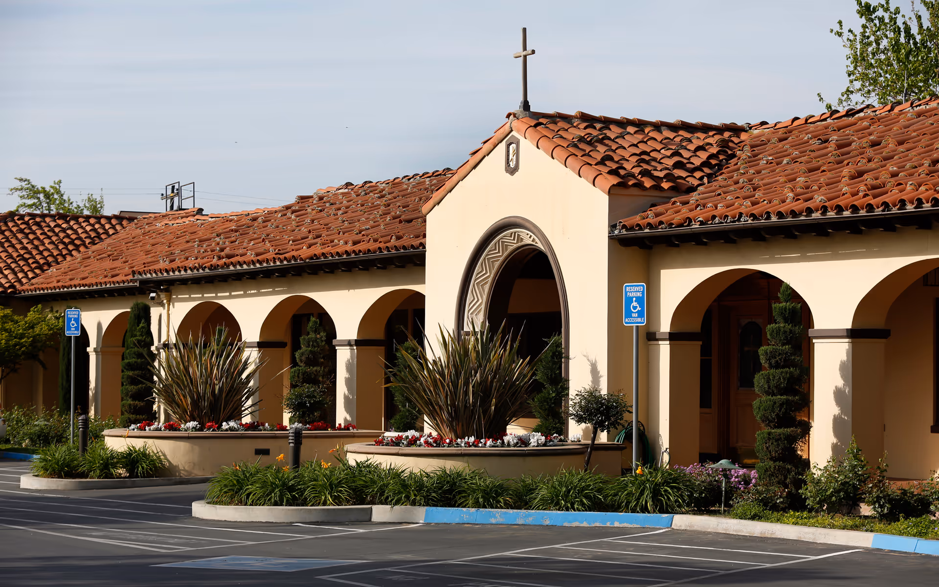 Exterior view of Vintage Gardens Assisted Living Community featuring a building with a terracotta tiled roof, arched walkways, a cross on top, landscaped flower beds, and reserved accessible parking spaces in front.