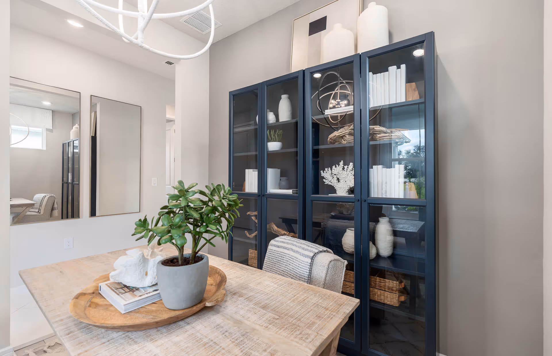 A modern dining area featuring a wooden table with a decorative tray holding a potted plant and books. Behind the table is a tall glass-front cabinet filled with white decorative items and books. Two large rectangular mirrors hang on the adjacent wall, reflecting part of the room.