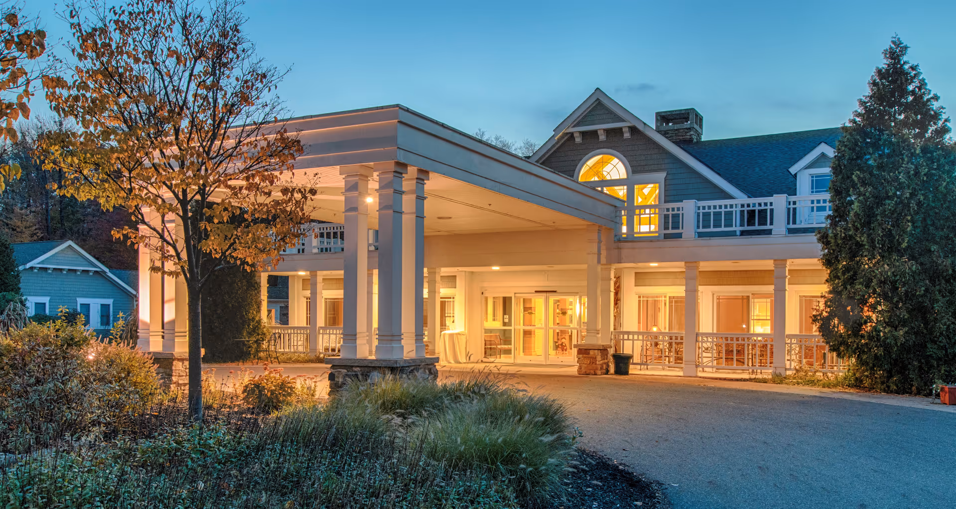 Exterior view of a senior living facility at dusk with warm lights glowing from inside. The building features a covered entrance supported by white columns, a balcony with white railings, and a peaked roof with large windows. Surrounding the entrance are landscaped bushes, a tree with autumn leaves, and a paved driveway.