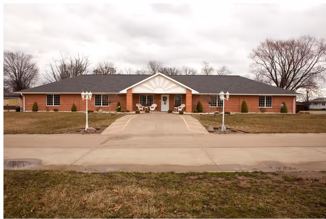 Front exterior view of a single-story brick building with a dark shingled roof, a central entrance with a small covered porch, two white lampposts along the driveway, and a lawn with small shrubs and trees in the background under a cloudy sky.