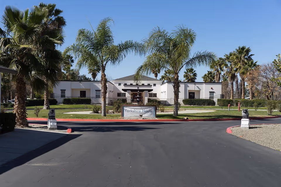 Exterior view of a single-story building named Yorkshire House, surrounded by palm trees and green lawns under a clear blue sky. The building has a white facade with a central entrance and a sign in front displaying the name.