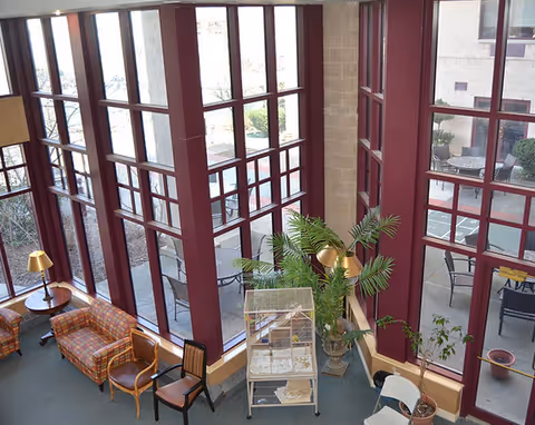 Interior view of a senior living facility lounge area with large floor-to-ceiling windows framed in dark red. The room contains several chairs and small tables, a birdcage on a stand, and potted plants. Outside the windows, there is a patio area with additional chairs and tables.