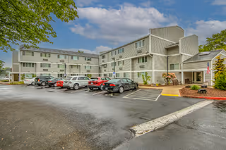 Exterior view of a three-story apartment building with a parking lot in front. Several cars are parked, and there are trees and landscaping around the building under a partly cloudy sky.