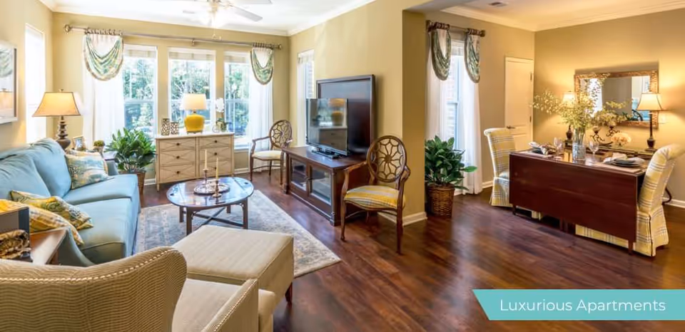 A bright and spacious living room with large windows letting in natural light. The room features a light blue sofa with decorative pillows, two beige armchairs, a round wooden coffee table, and a TV on a wooden stand. There are plants and lamps adding to the cozy atmosphere. Adjacent to the living area is a dining space with a dark wooden table, four yellow plaid chairs, a large mirror on the wall, and decorative items on the table. The floor is wooden, and the walls are painted a warm beige color.