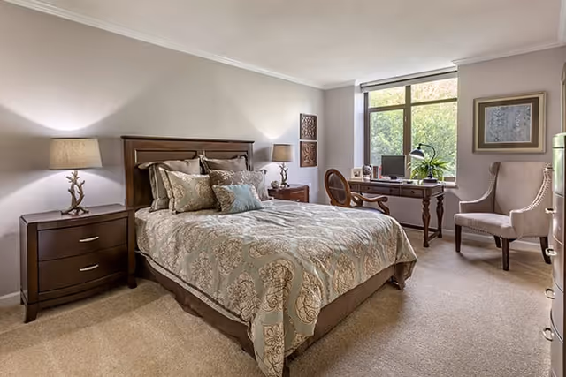 A well-lit bedroom featuring a large bed with patterned bedding and multiple pillows, two wooden nightstands each with a lamp, a wooden desk with a computer and a plant, a wooden chair, a cushioned armchair, and a large window letting in natural light.