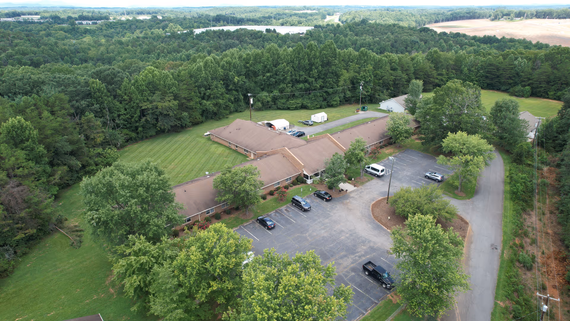 Aerial view of Yadkin Valley Senior Living facility surrounded by trees and greenery. The building has a brown roof and is shaped in a U-formation with a parking lot in front containing several cars. The area around the building is lush with grass and trees, and there are hills and more trees visible in the background.
