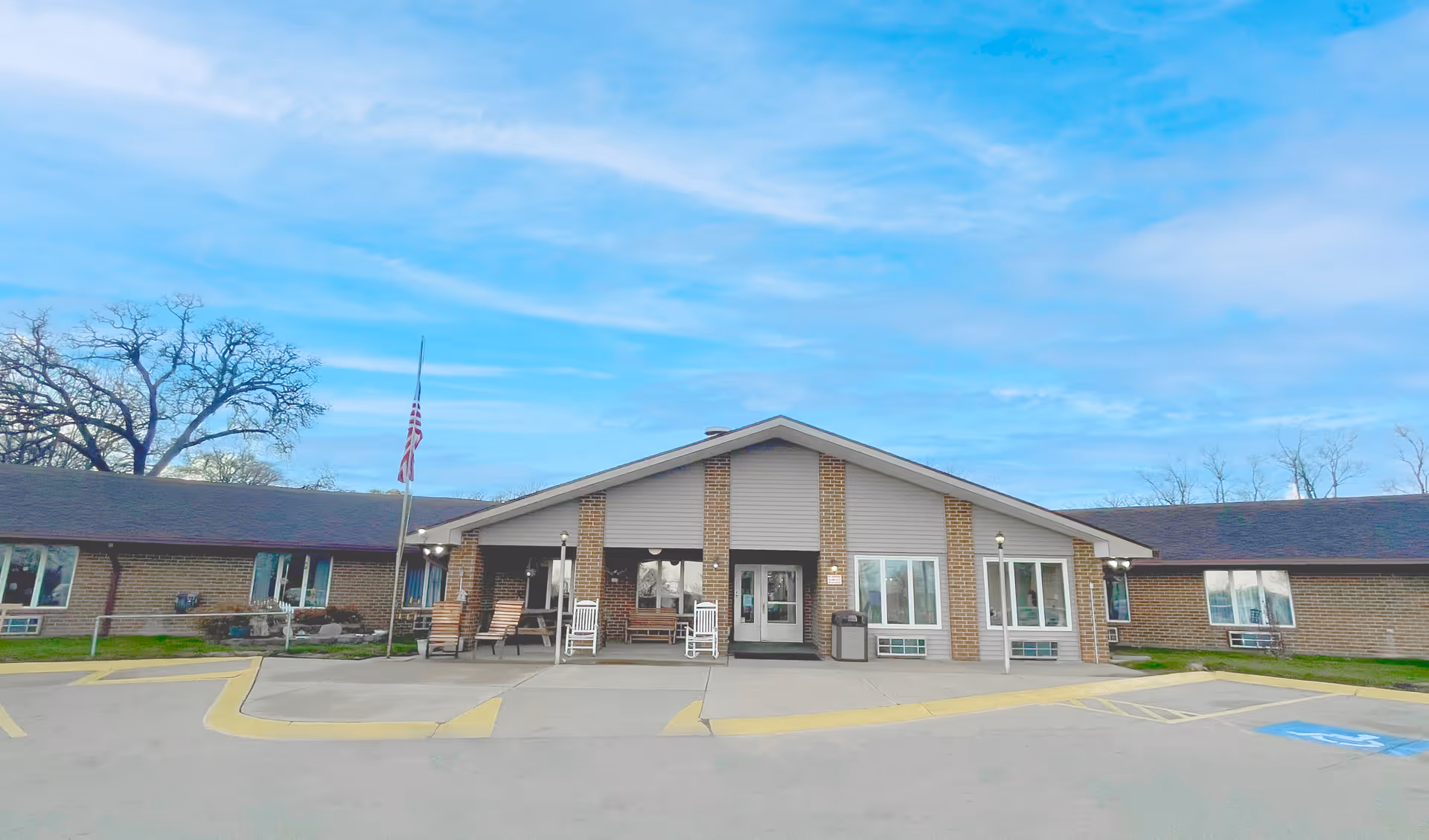 Front exterior view of Maryville Living Center, a single-story building with a peaked roof, brick and siding facade, several windows, a covered entrance with rocking chairs, an American flag on a flagpole, and a parking area with handicap spaces in front.