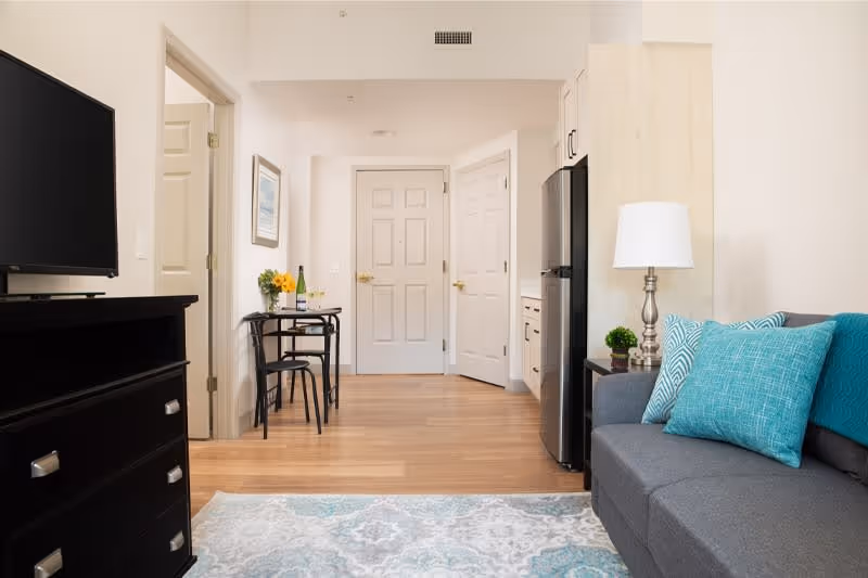 Interior view of a senior living facility apartment showing a small living area with a gray couch adorned with blue and turquoise pillows, a side table with a lamp and small plant, a black dresser with a flat-screen TV, and a small dining table with two chairs near the entrance door. The floor is wooden with a patterned area rug in front of the couch.