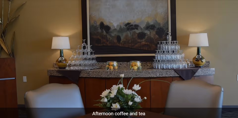 A cozy interior space with a granite countertop holding two pyramid stacks of upside-down glasses, two glass bowls filled with lemons and oranges, and two table lamps with white shades. A large framed painting of trees hangs on the wall behind the countertop. In the foreground, there is a table with a floral centerpiece and two chairs.