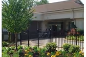 Exterior view of Sunny Hill Nursing Home-Will County showing the entrance area with a covered drop-off zone, surrounded by a black metal fence and landscaped with green shrubs and yellow flowers. A tree is visible on the left side of the image.