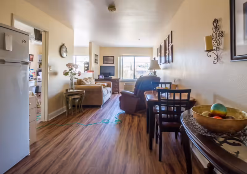 Interior view of a senior living facility apartment featuring a living room with a sofa, armchair, TV on a wooden stand, and a dining area with a small table and chairs. The room has wooden flooring, beige walls, and decorative wall art. A refrigerator is visible on the left side near the entrance to another room.