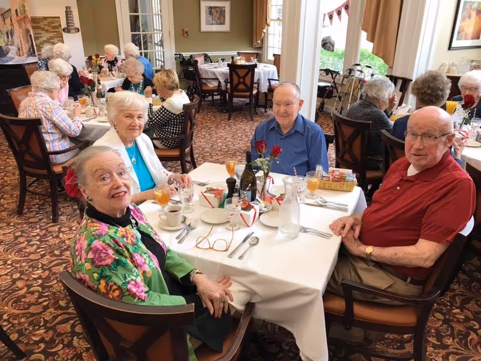 A group of elderly people sitting around a table in a dining room at a senior living facility. The table is set with plates, cups, glasses of iced tea, and a vase with red roses. The room has carpeted floors, multiple tables with other elderly residents dining, and large windows letting in natural light.
