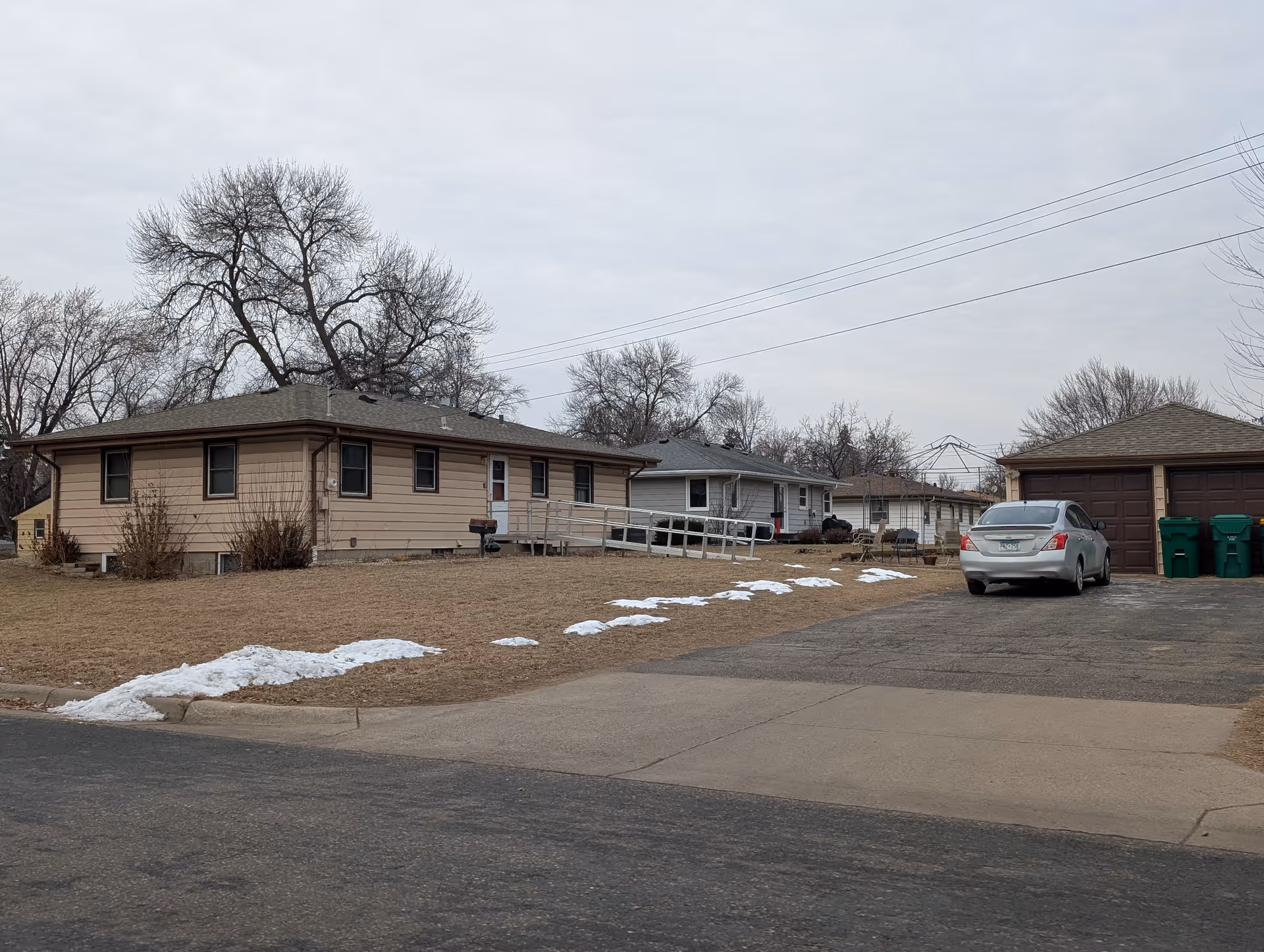 Exterior view of a single-story residential building with beige siding and a wheelchair ramp leading to the front door. There is a driveway with a silver car parked next to a two-car garage. Leafless trees and patches of snow are visible on the brown grass under an overcast sky.