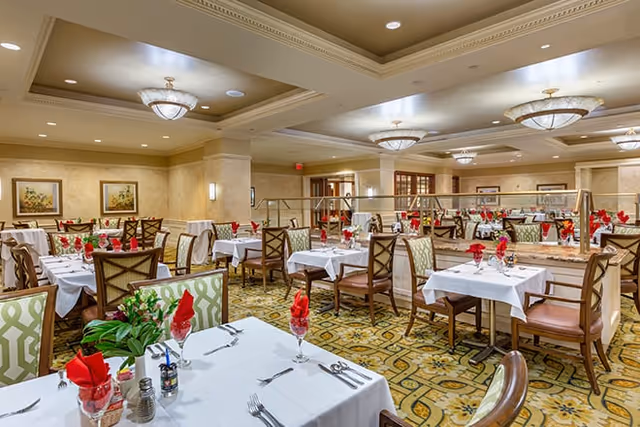 A spacious dining room with multiple tables covered in white tablecloths, each set with silverware, glasses, and red folded napkins. The room features patterned carpet, cushioned chairs with wooden frames, warm lighting from ceiling fixtures, and framed artwork on the walls.