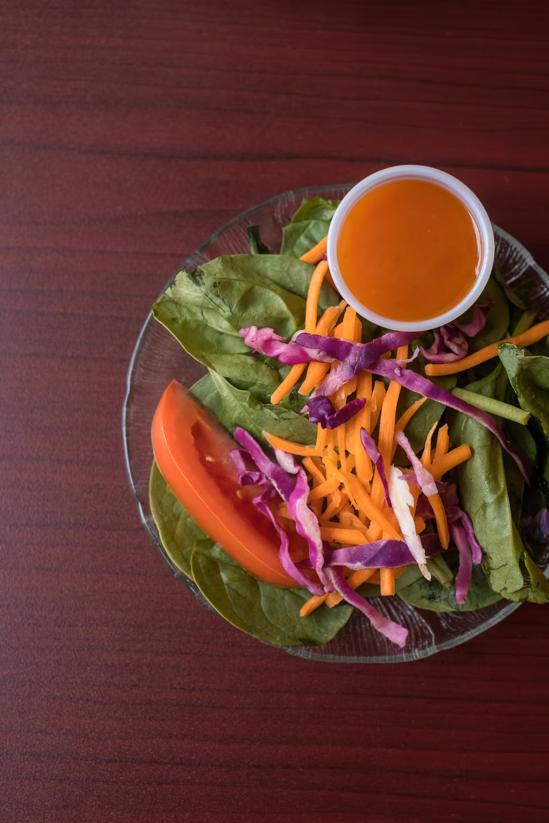 A clear glass plate with a fresh salad consisting of spinach leaves, shredded carrots, shredded purple cabbage, and a slice of tomato, accompanied by a small cup of orange salad dressing on a dark wooden table.