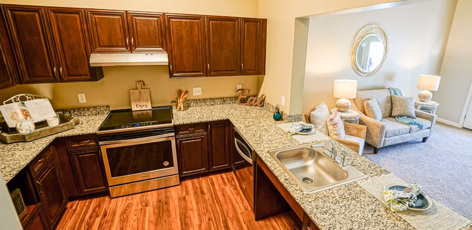 A modern kitchen with dark wooden cabinets, granite countertops, a stainless steel oven, and a sink. The kitchen opens into a living area with a beige sofa, armchair, two lamps, and a round mirror on the wall.