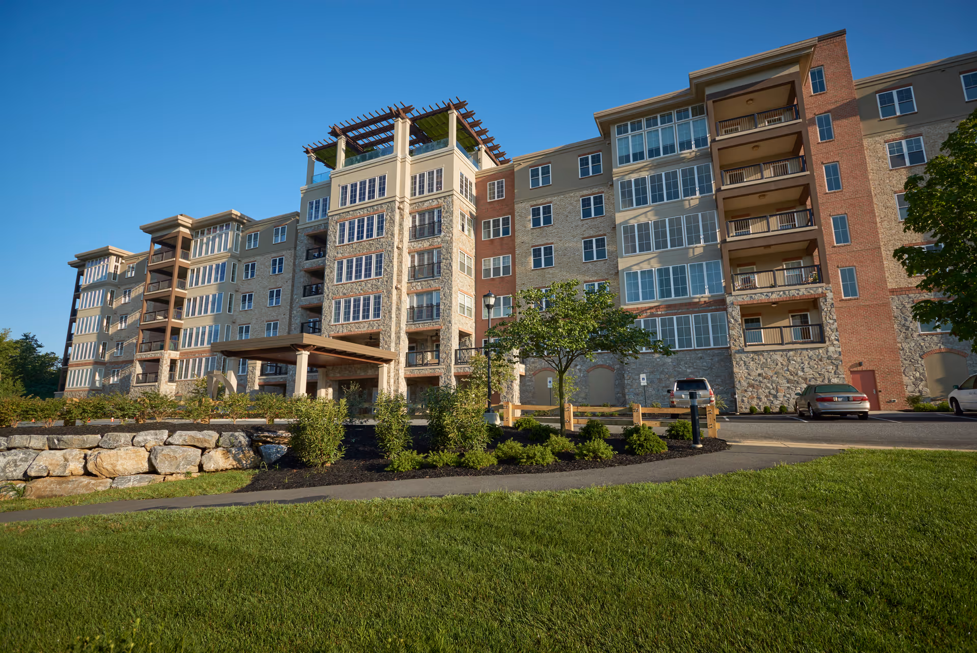 Exterior view of a multi-story senior living facility building with balconies, large windows, and a covered entrance. The foreground features a well-maintained lawn, shrubs, and a stone retaining wall under a clear blue sky.