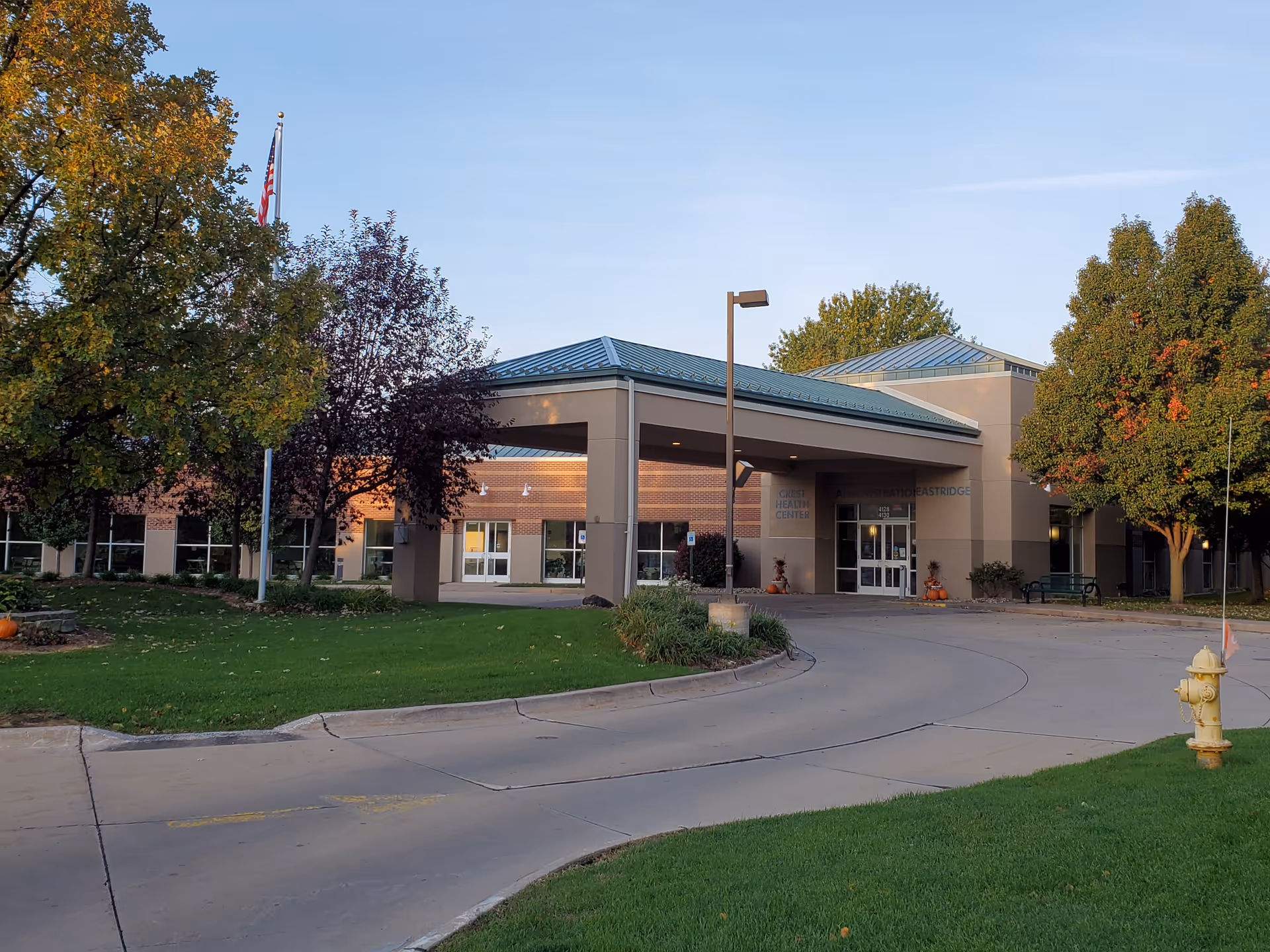 Exterior view of a single-story building with a covered entrance and a curved driveway. The building is surrounded by green grass, trees with autumn foliage, and a yellow fire hydrant near the driveway. The sky is clear and blue.