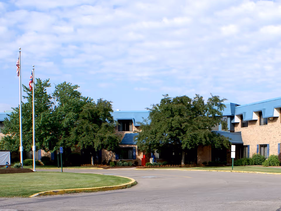 Exterior view of Lake Pointe Health Center building with a blue roof, beige brick walls, and several windows. There are two flagpoles with the American flag and another flag, green trees, and a curved driveway in front of the building under a partly cloudy sky.