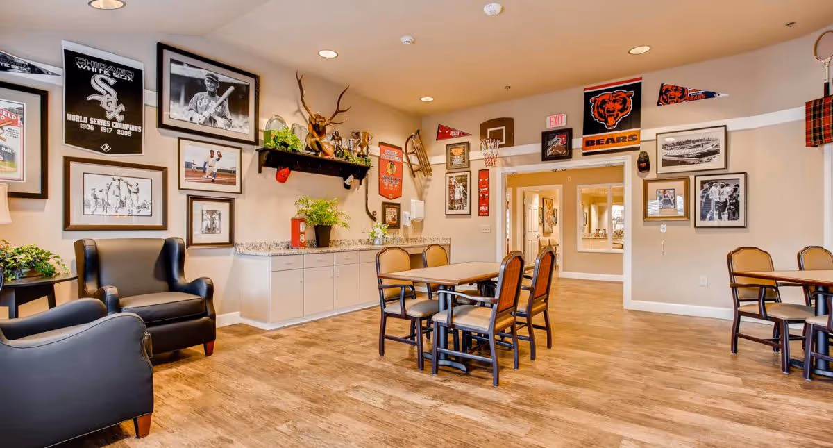 A cozy common area in a senior living facility decorated with sports memorabilia including framed photos, banners, and trophies. The room features wooden flooring, two black armchairs, tables with chairs, and a countertop with plants and decorations. The walls are beige, and the lighting is warm with recessed ceiling lights.