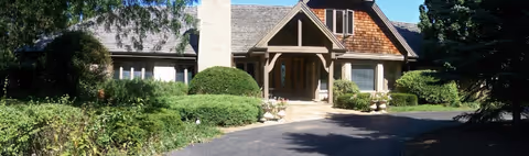 Front exterior view of a single-story residential building with a shingled roof, a chimney, and a covered entrance supported by wooden beams. The building is surrounded by well-maintained bushes, trees, and a paved driveway.