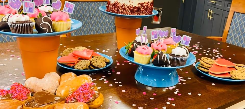 A wooden table decorated with pink and white confetti and star-shaped sprinkles, holding multiple blue plates and stands with various desserts including cupcakes with pink and white frosting topped with small heart and 'LOVE' signs, chocolate-covered strawberries, heart-shaped red cookies, chocolate chip cookies, donuts with pink sprinkles, and a cake on a stand in the background. The setting appears to be a dining area with patterned chairs and cabinets in the background.