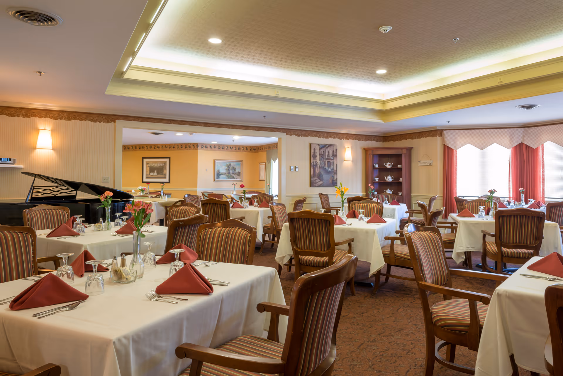 A well-lit dining room with multiple tables covered in white tablecloths, each set with red folded napkins, glassware, and silverware. The room features striped upholstered wooden chairs, a grand piano in the corner, floral centerpieces on the tables, and warm yellow walls with framed artwork. Large windows with red curtains allow natural light to enter the space.