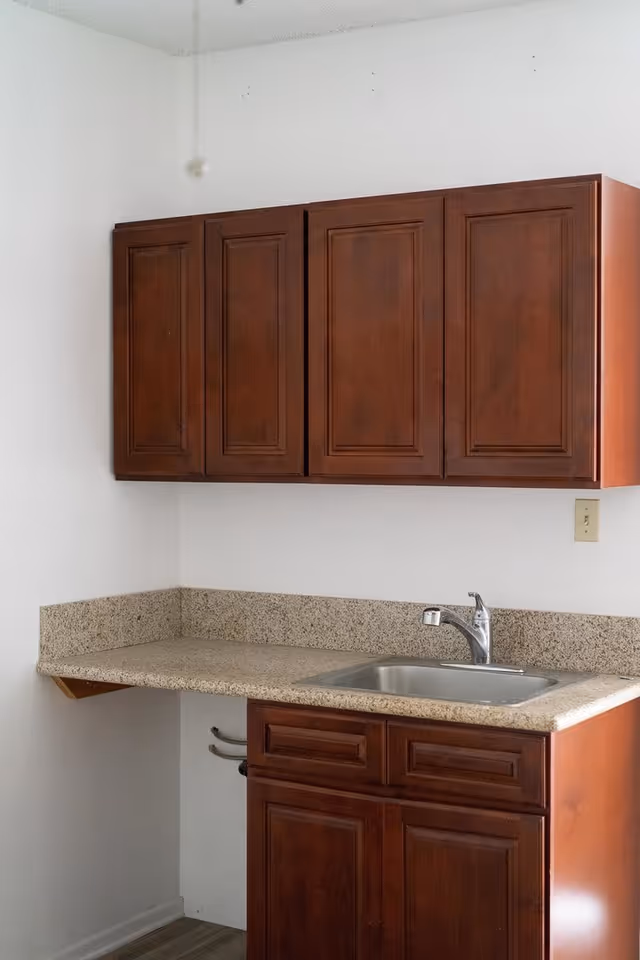 A small kitchen area with wooden cabinets, a granite countertop, and a stainless steel sink with a faucet. The walls are white and there is a light switch on the right side.