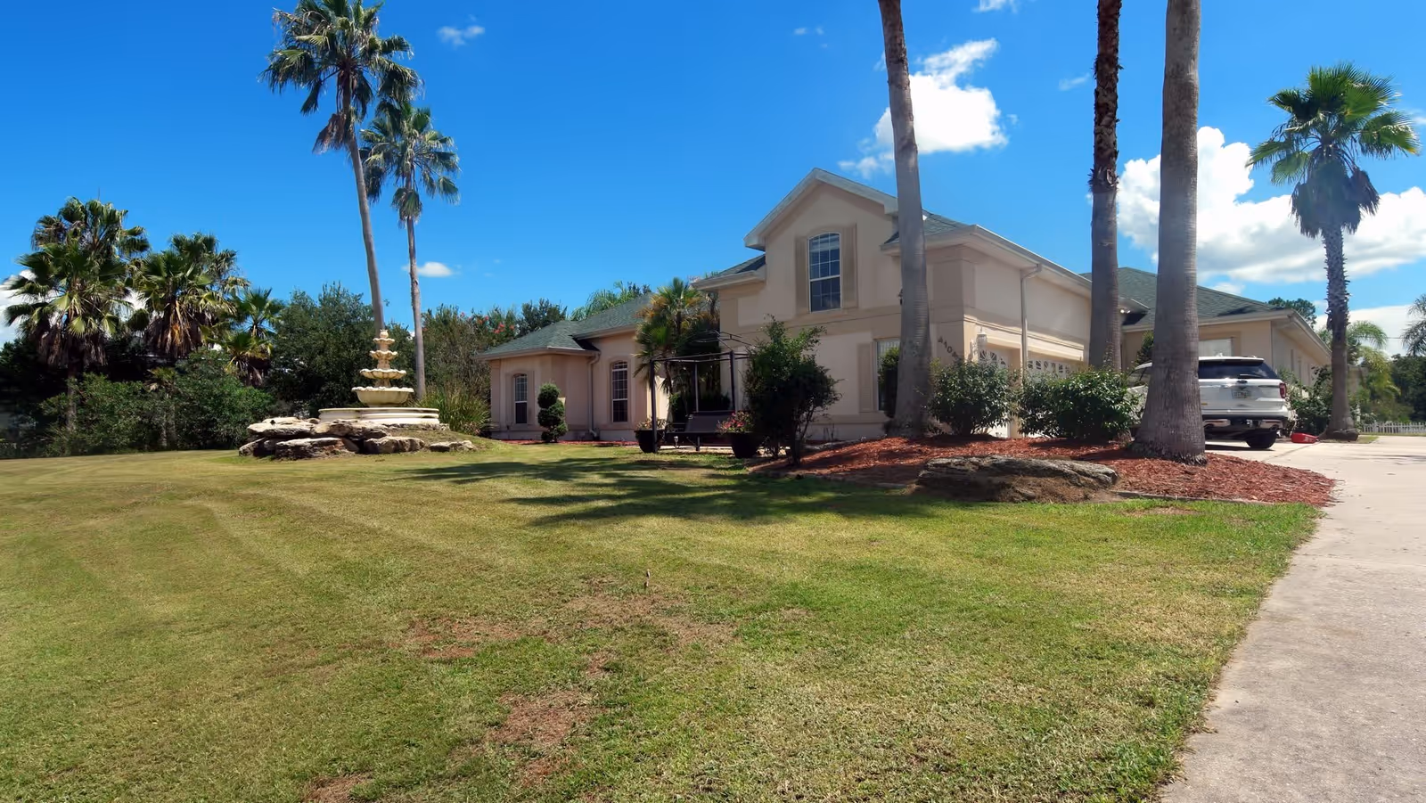 Exterior view of a single-story residential building with a green roof, surrounded by palm trees and a well-maintained lawn. There is a multi-tiered fountain on the lawn and a driveway with a white SUV parked near the building. The sky is clear with a few clouds.