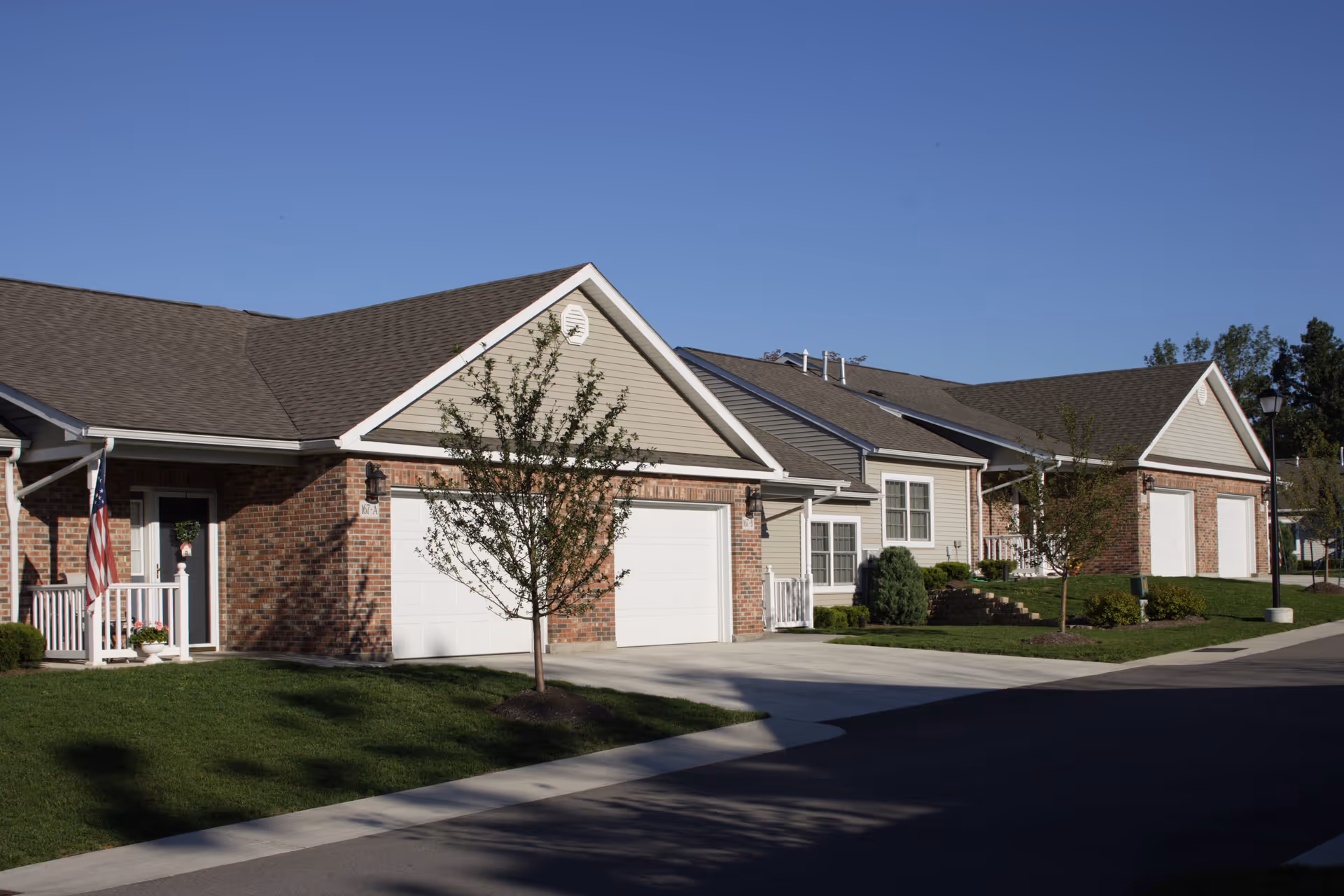 Exterior view of a residential senior living facility with brick and siding buildings, garages, small trees, and a well-maintained lawn under a clear blue sky.