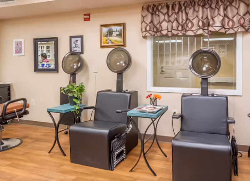 Interior of a senior living facility hair salon with two black salon chairs under hair dryers, small side tables with magazines and a plant, framed artwork on the wall, and a window with patterned curtains.