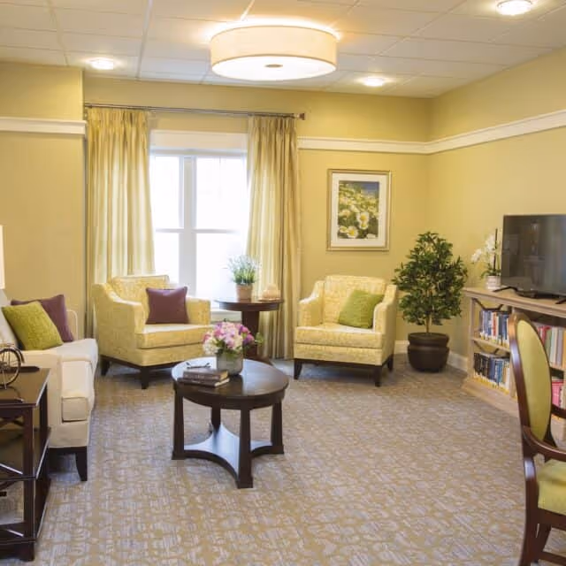 A cozy living room with two yellow armchairs and a white sofa, each with colorful throw pillows. A round wooden coffee table with a flower arrangement and books sits in the center. There is a window with yellow curtains, a framed floral picture on the wall, a potted plant, and a TV on a wooden stand with bookshelves underneath. The room has beige walls and carpeted floor.