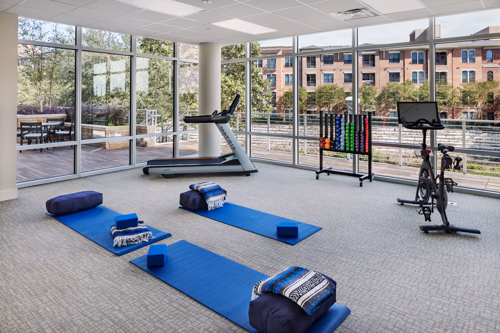 A bright exercise room with large floor-to-ceiling windows showing an outdoor patio and brick buildings outside. Inside, there are three blue yoga mats each with a bolster, blanket, and yoga blocks. The room also contains a treadmill, a stationary bike, and a rack with colorful dumbbells.