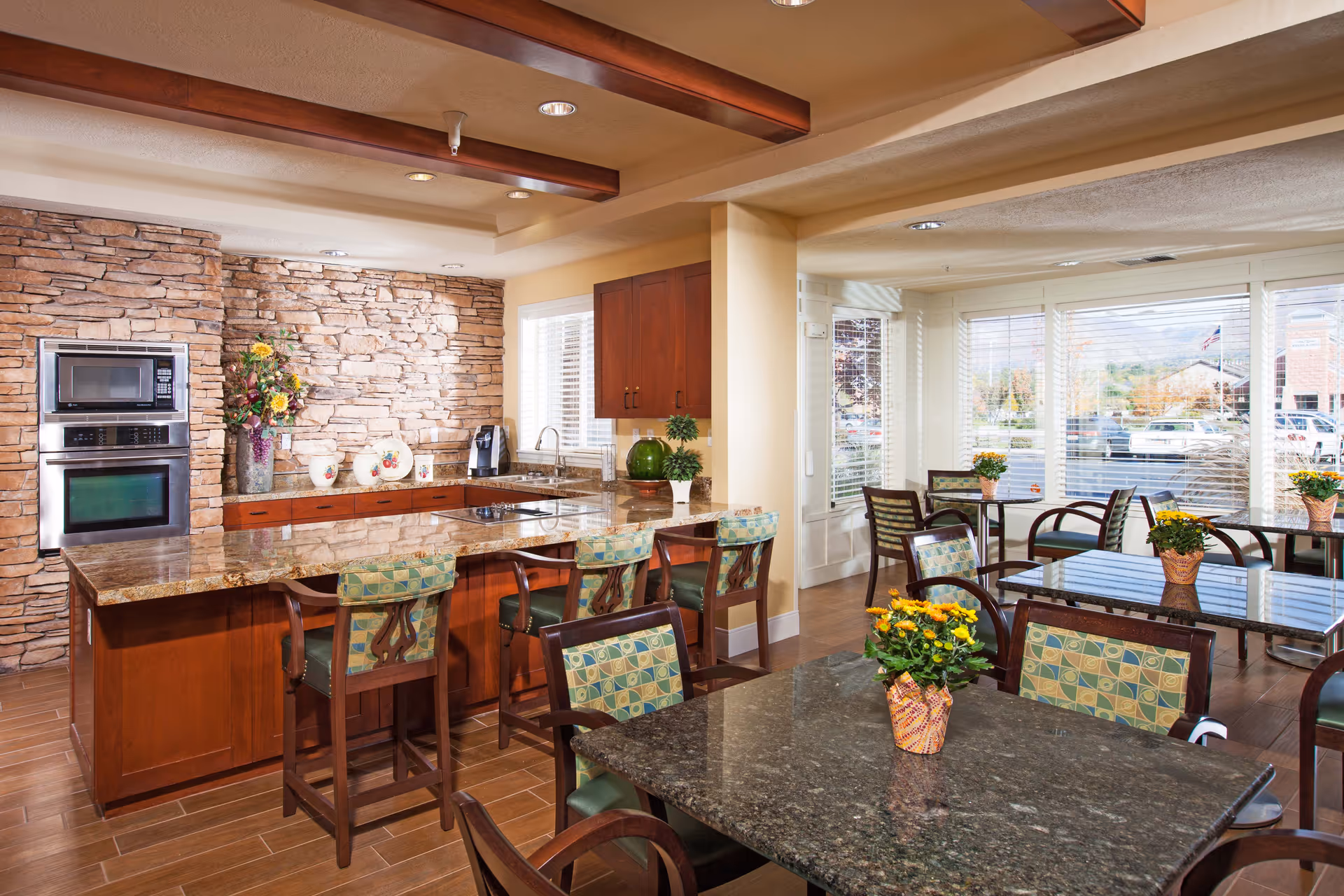 A bright and inviting dining area with multiple tables and chairs, each table decorated with a small potted plant with yellow flowers. The room features large windows with white blinds allowing natural light to fill the space. Adjacent to the dining area is a kitchen with a stone accent wall, built-in oven and microwave, wooden cabinets, and a granite countertop island with bar stools.