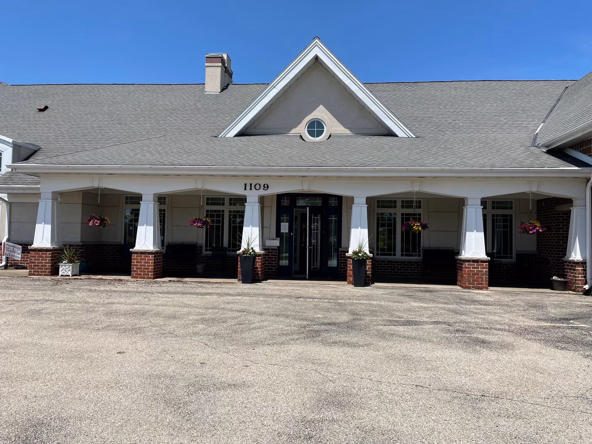 Front exterior view of a single-story building with a gray shingled roof, white columns, brick accents, and hanging flower baskets. The building has the number 1109 above the entrance doors and is under a clear blue sky.