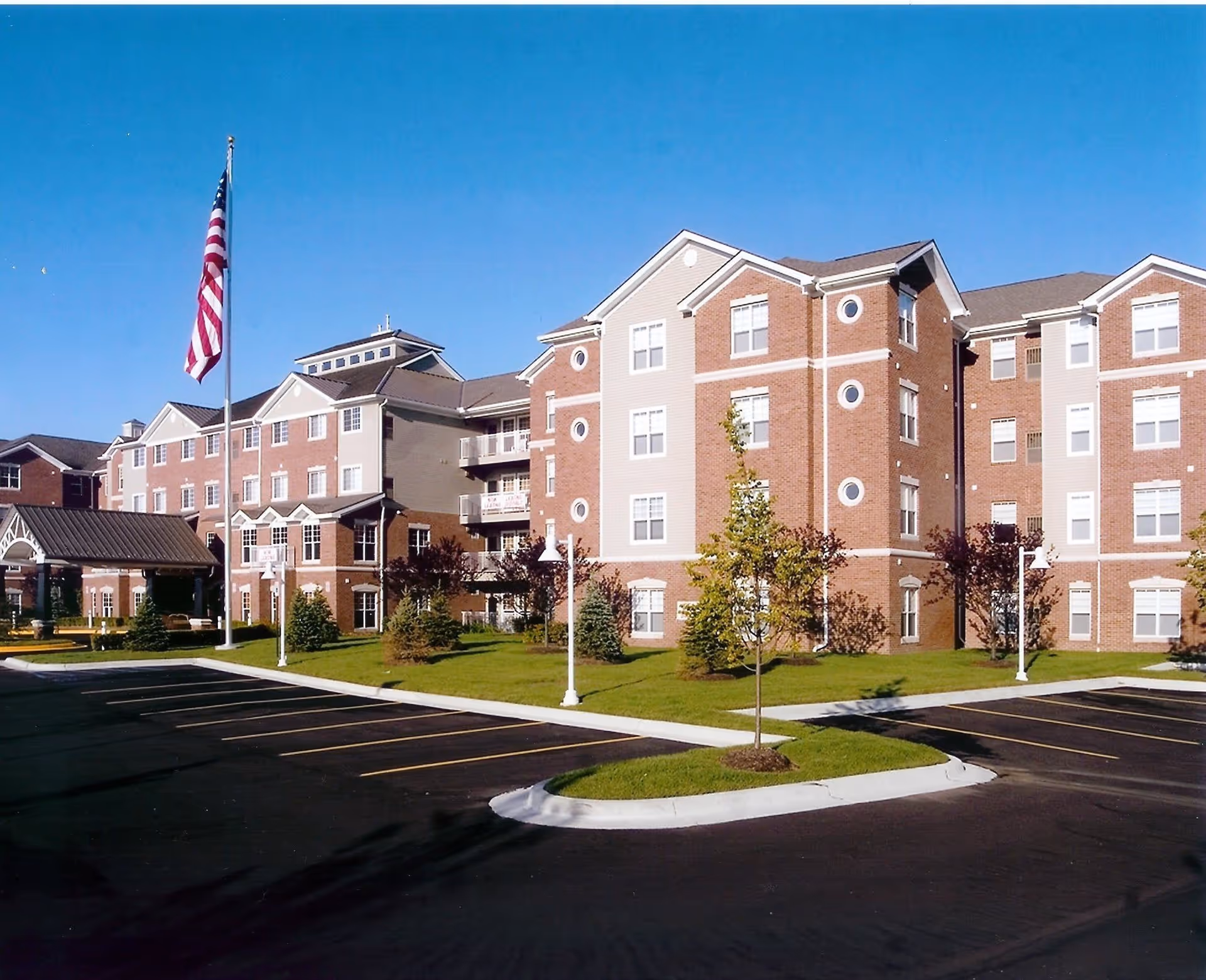 Exterior view of a multi-story senior living facility building with brick and beige siding, surrounded by a well-maintained lawn, small trees, and a parking lot. An American flag is flying on a flagpole near the entrance under a covered driveway.