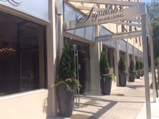 Exterior view of the entrance to Signature Senior Living facility with large windows and a metal awning displaying the facility name. Several tall potted plants line the walkway leading to the entrance.
