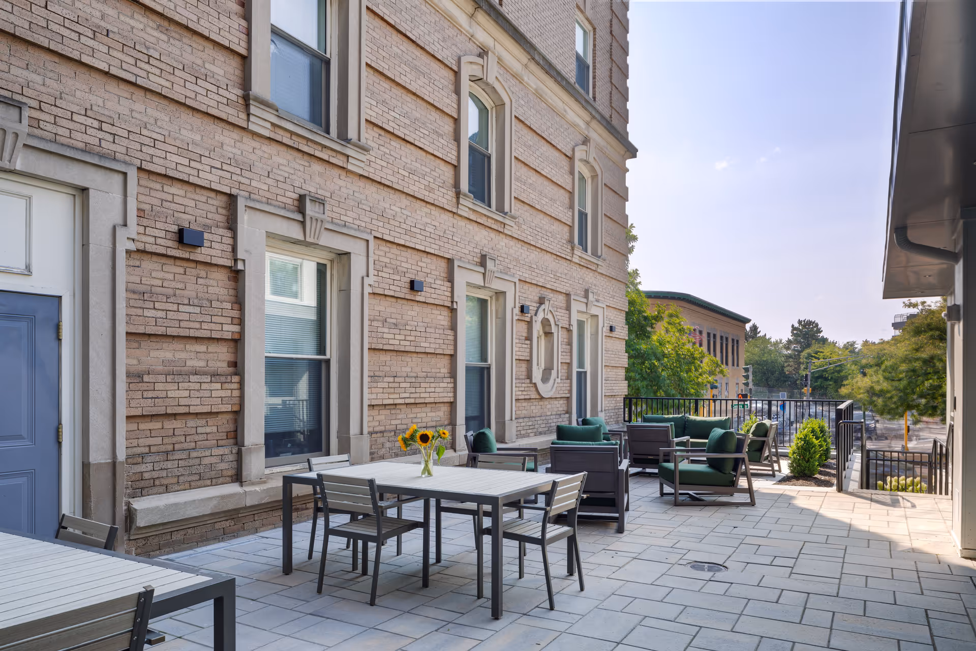 Outdoor patio area at Evans Park at Newton Corner with tables and chairs, some with green cushions, against a brick building. There is a vase with sunflowers on one of the tables and trees and a street visible in the background under a clear sky.