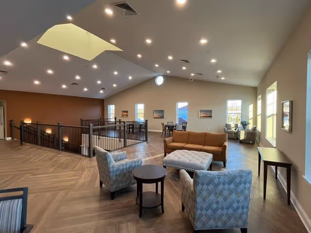 A spacious, well-lit common area with high vaulted ceilings and recessed lighting. The room features a seating arrangement with a brown sofa, two patterned armchairs, and a large tufted ottoman in the center. There are several windows allowing natural light to fill the space, and a railing encloses a stairwell area. Additional seating and tables are visible in the background near the windows.