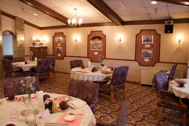 A cozy dining room with round tables covered in white lace tablecloths, each set with pink placemats, cups, napkins, and condiments. The chairs have a floral upholstery. The walls are decorated with framed pictures and sconces, and the ceiling has wooden beams and a chandelier. The carpet has a patterned design.