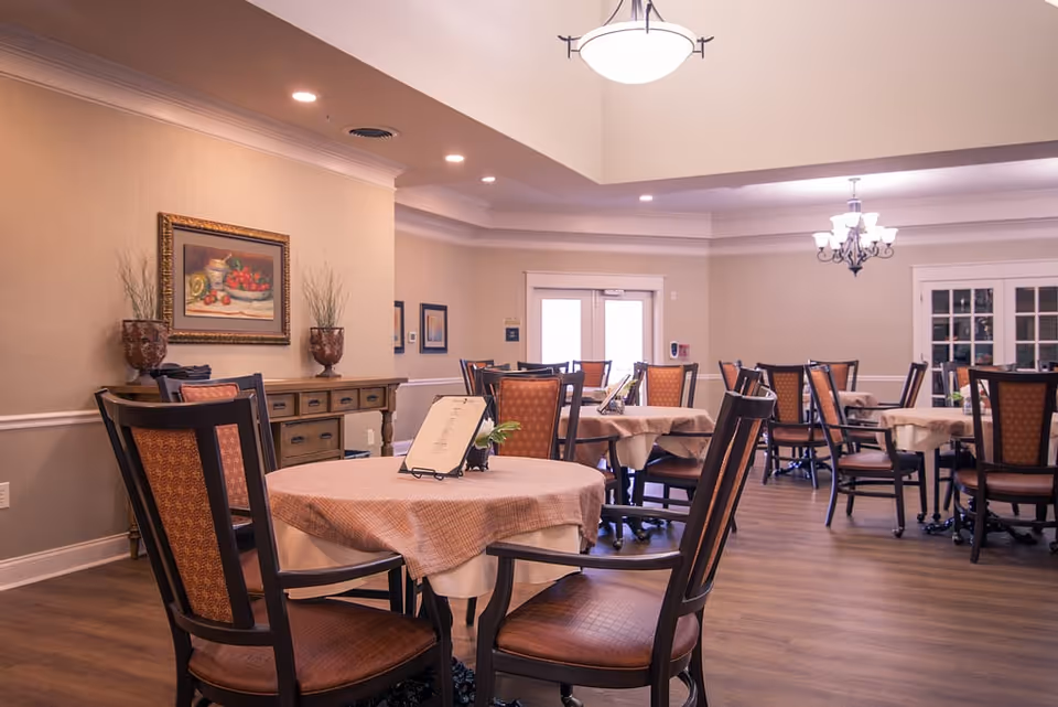 A dining room in a senior living facility with multiple round tables covered with beige tablecloths and surrounded by wooden chairs with cushioned seats and backs. The room has warm lighting from ceiling fixtures, a wooden sideboard with decorative vases and a framed painting on the wall, and double doors with glass panels in the background.