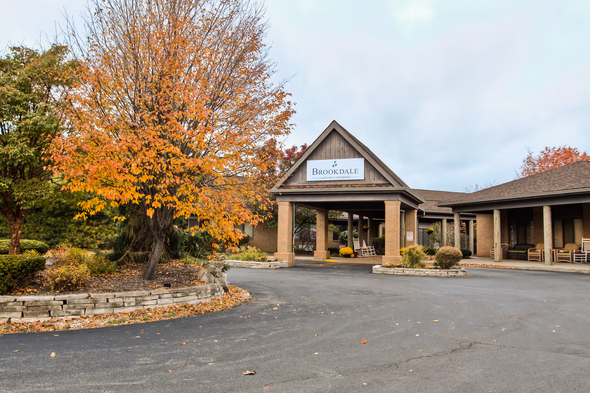 Exterior view of Brookdale Lakeview Crossing facility entrance with a covered driveway, surrounded by autumn trees with orange and yellow leaves, and landscaped garden beds.